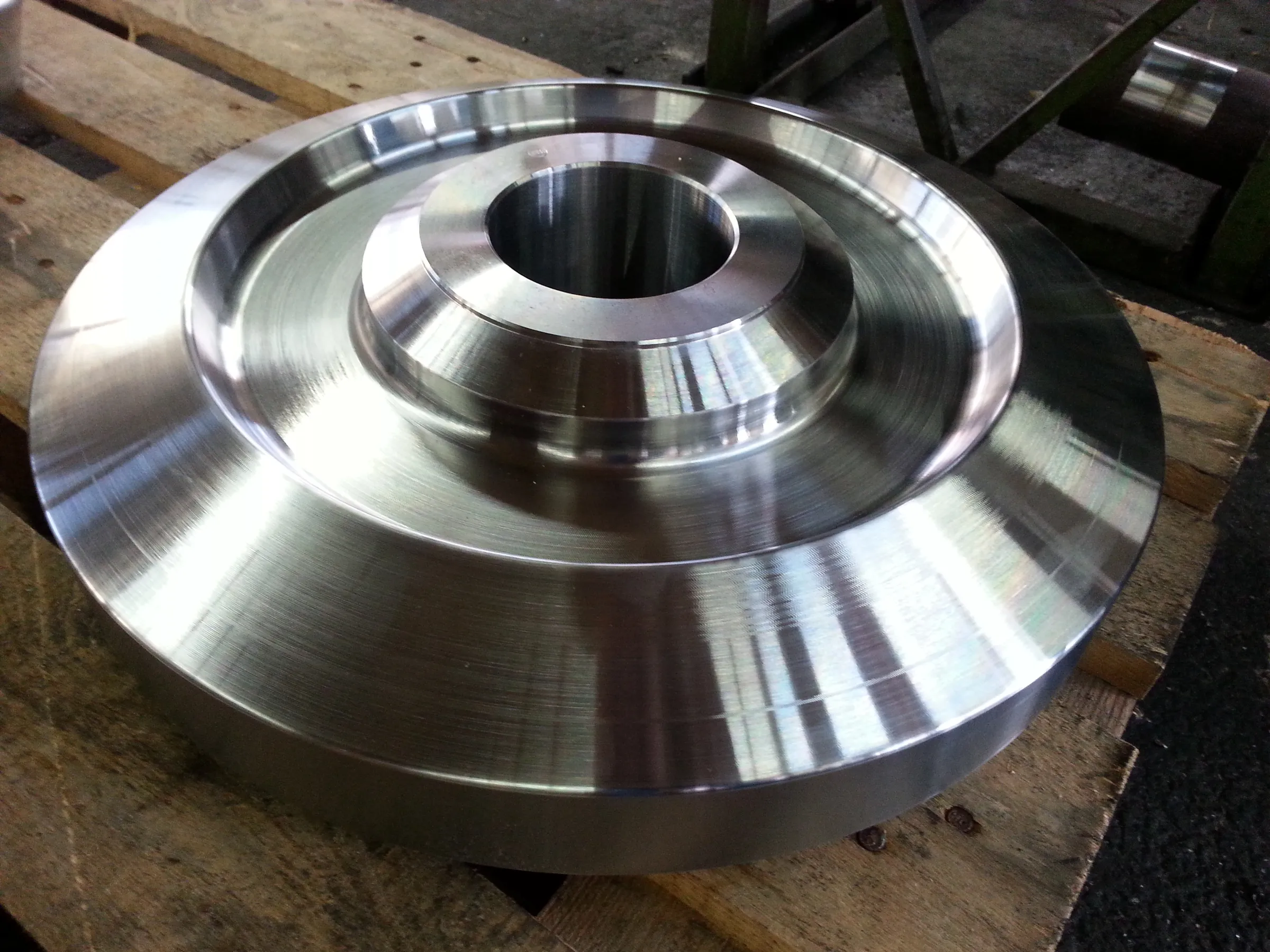 Großes Drehteil aus Metall: Scheibe mit abgesetzter Nabe und Innenbohrung, mit feinen Drehriefen, auf einer Holzpalette.