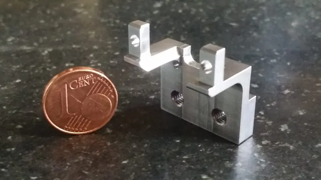 Kleines Frästeil aus Metall mit zwei Laschen und Gewindebohrungen, daneben eine 1-Cent-Münze zum Größenvergleich.
